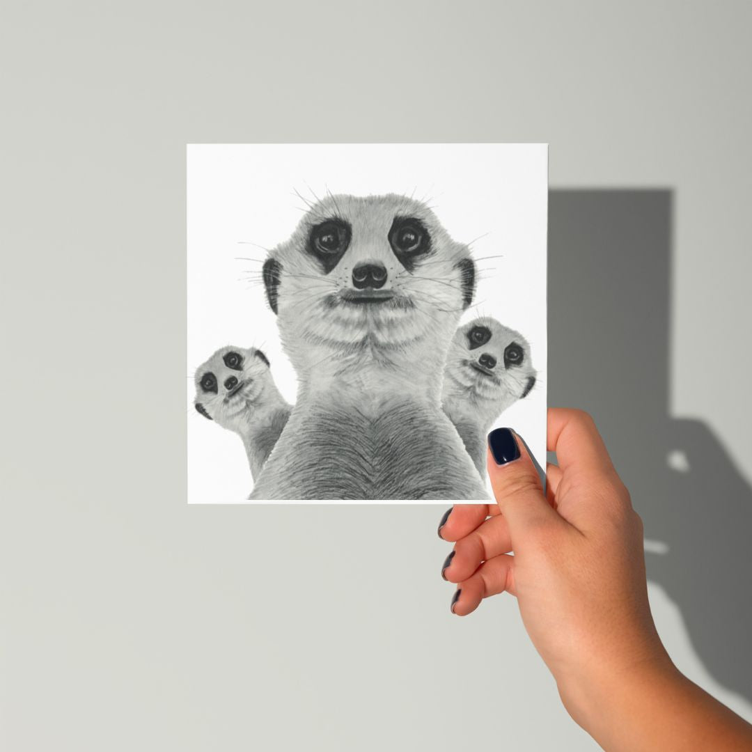 Meerkat Madness Greeting CardA hand holding the Meerkat Madness Greeting Card, showcasing a fun, hand-drawn illustration of a meerkat family in humorous poses. The card is held upright, displaying the lively design.